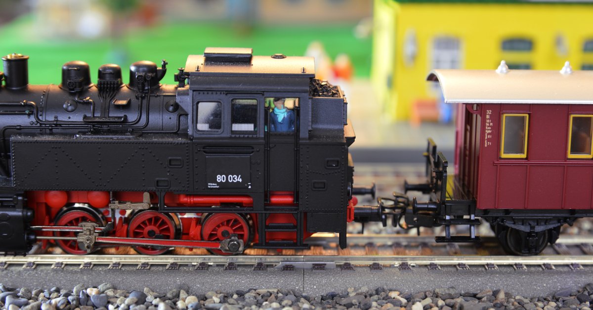 A black and red model train connected to a red and yellow train car sits on gravel tracks beside a yellow building.